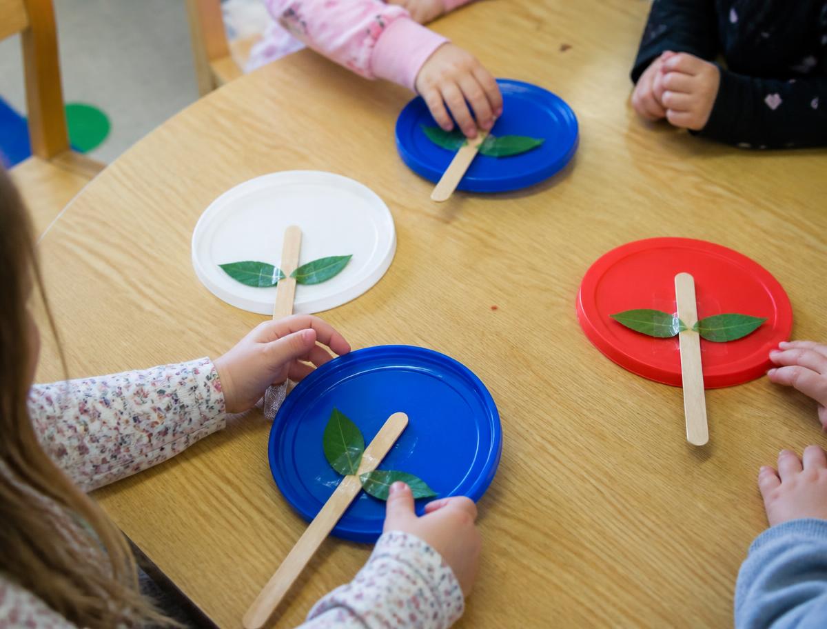 Children using large plastic ice cream container lids and wooden sticks to make simple flowers at a timber table