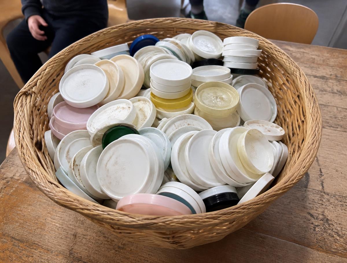 A woven basket of well-used yoghurt container lids collected over time, different brands, sizes and shades, on a timber classroom table
